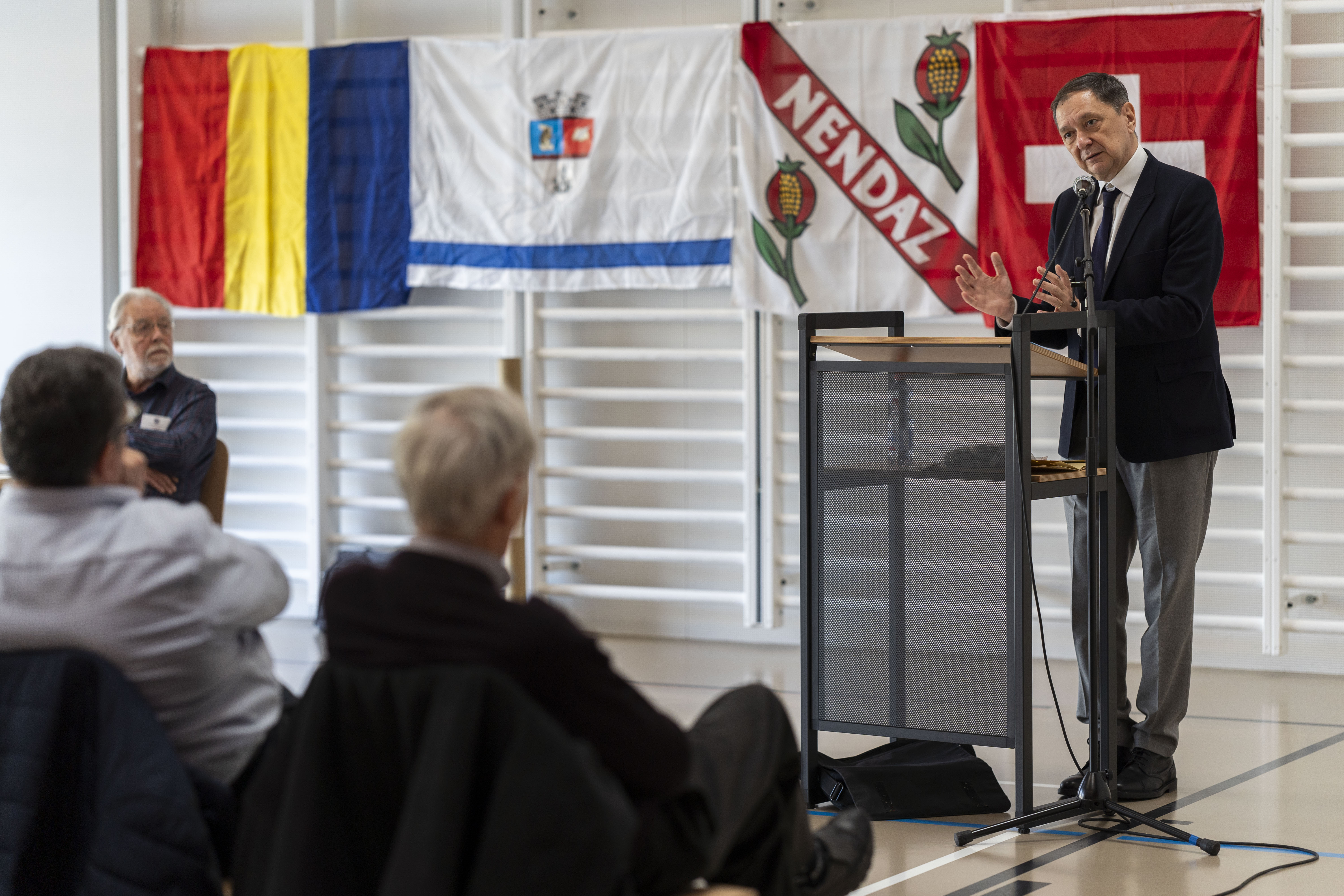 Person auf dem Podium, die sich an ein Publikum wendet, mit Fahnen im Hintergrund.