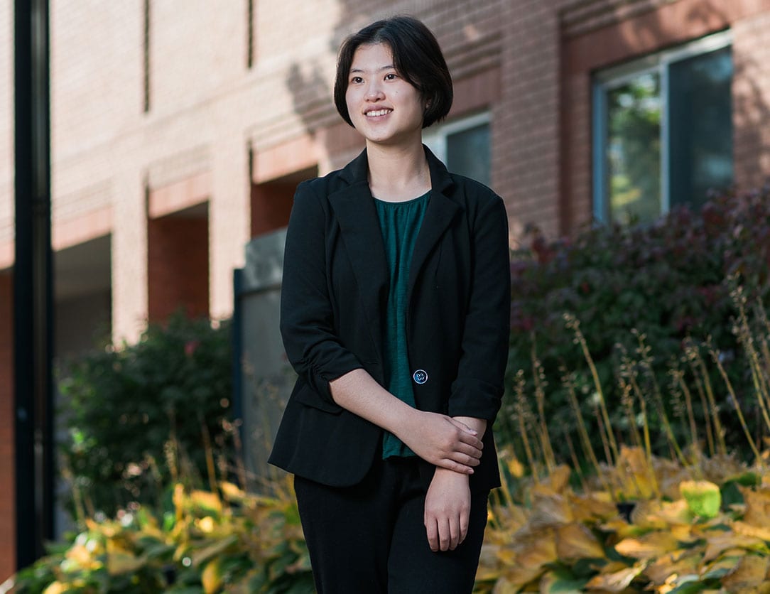 Image of Alice Lo standing outside of the U of T Innis building, wearing a green shirt, black blazer and pants.