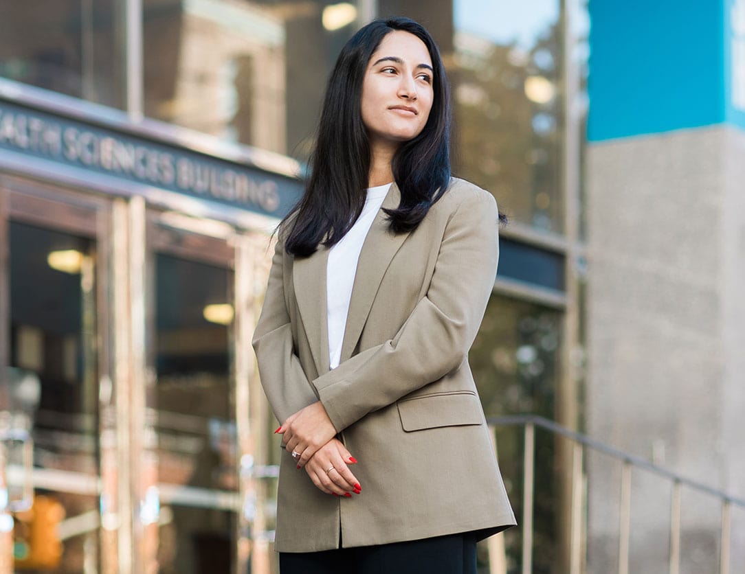 Image of Shakila Khalkhali Ardestani standing outside of the U of T Nursing building, wearing a white shirt and tan blazer.