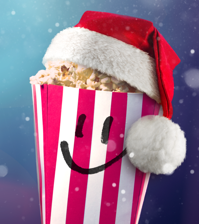 Ein gefüllter Popcorn-Becher mit Smiley-Gesicht und Weihnachtsmütze vor einem leeren Kinosaal. Im Hintergrund blendet das Licht des Filmprojektors.