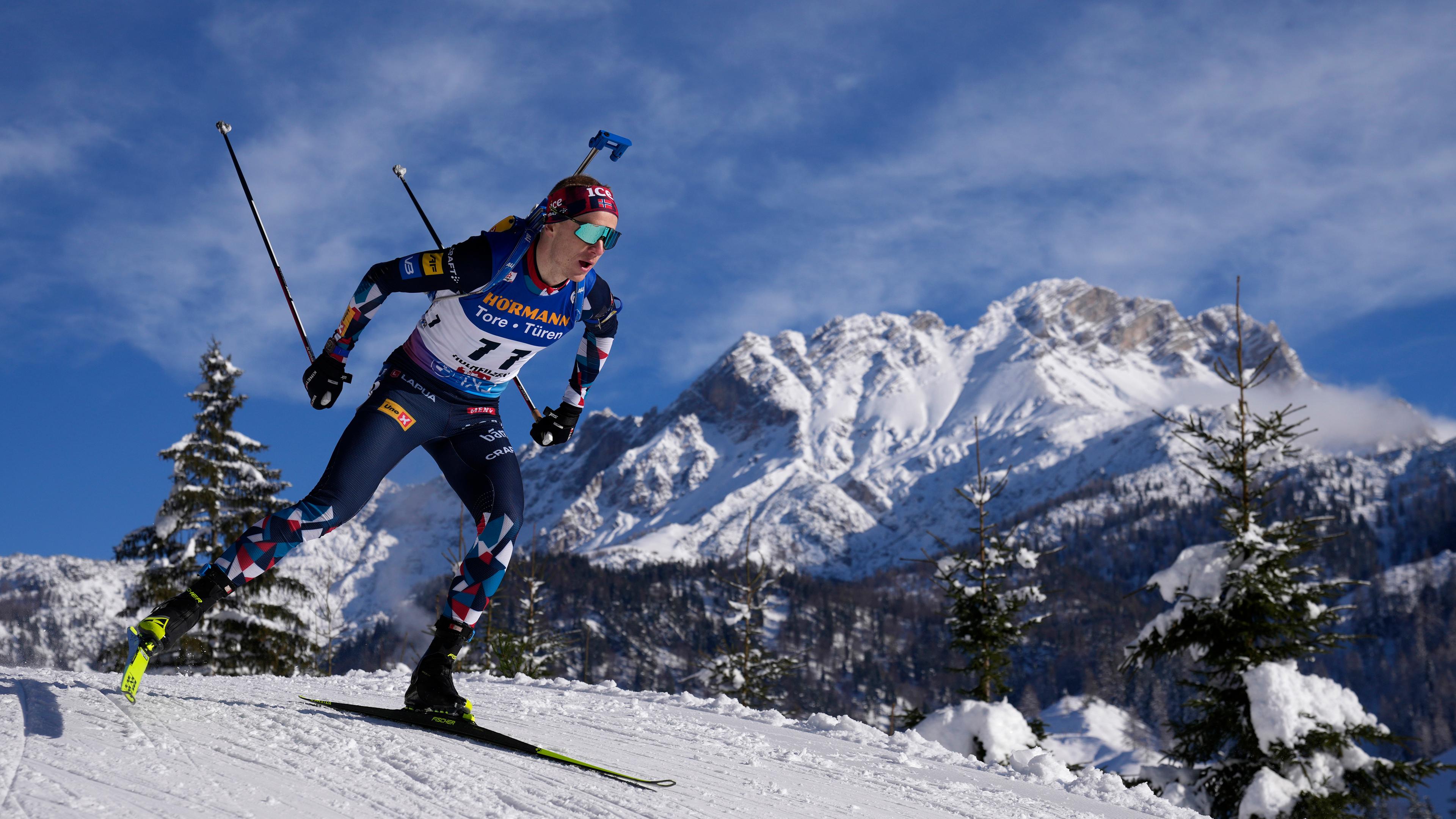 Ein Biathlon-Sportler auf einer Piste mit verschneiten Bergen im Hintergrund.