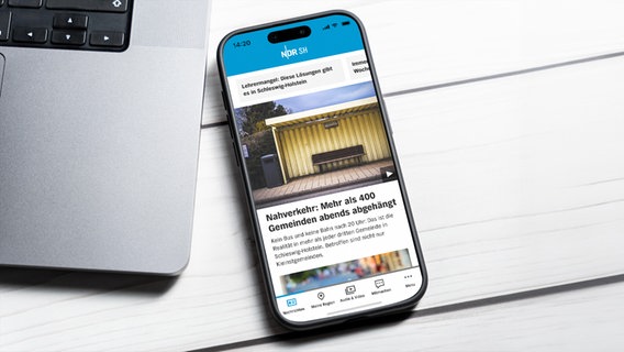 Ein Smartphone auf dem die NDR SH App zu sehen ist, liegt auf dem Tisch neben einem Laptop. © IMAGO Foto: Bihlmayerfotografie