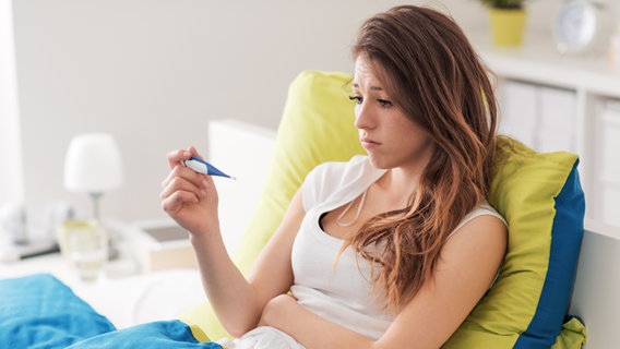 Eine junge Frau sitzt aufrecht in einem Bett und schaut betrübt auf ein Fieberthermometer, das sie in der Hand hält © colourbox Foto: Andrea De Martin