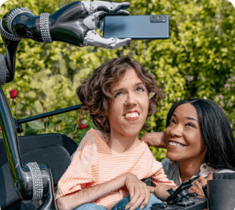 A white person with wavy brown hair sits in their motorized wheelchair with a robot arm attachment holding an Android phone. A Black woman crouches next to them and smiles up at the Android phone.