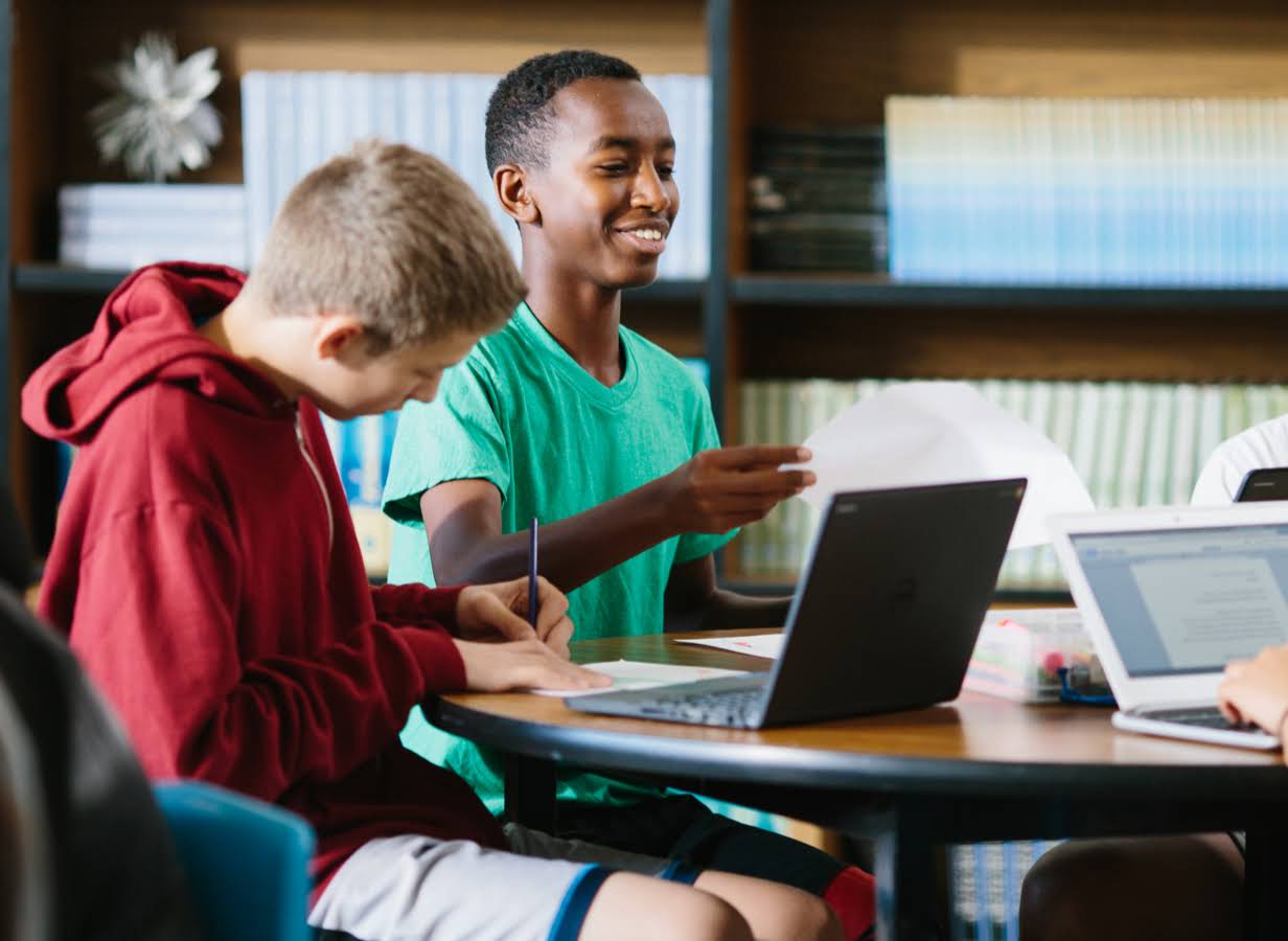 Zwei Schüler; der eine hält ein Blatt Papier in der Hand, der andere arbeitet an einem Chromebook