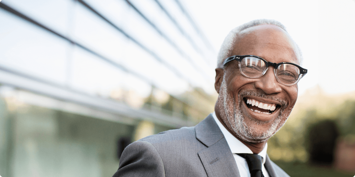 A cheerful elderly man with glasses and in a grey suit, outside an office building.