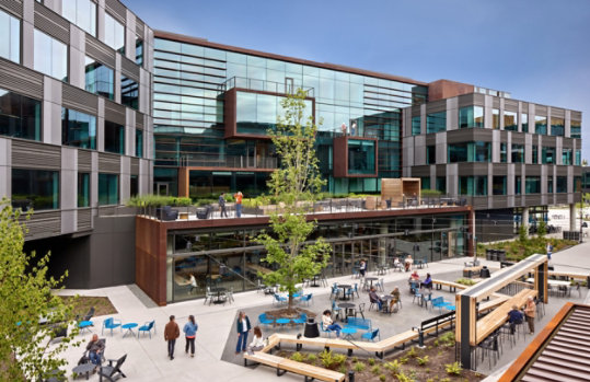 An exterior view of Microsoft office buildings in Redmond, Washington, showing people walking around a plaza amongst wooden benches, blue patio furniture and trees