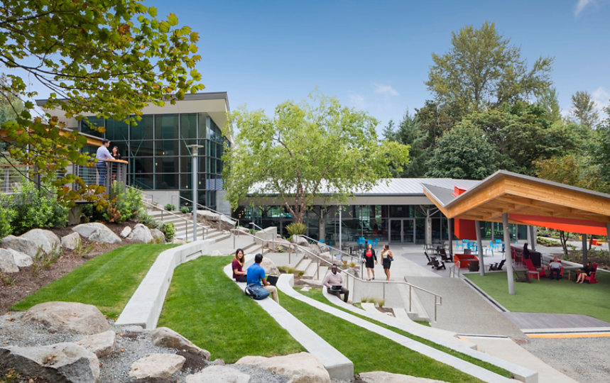 A view of several small groups of adults standing, sitting on steps or in chairs, and walking between low office buildings among trees and with a blue sky overhead.