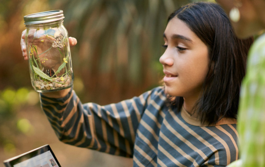 A child looks inside a glass jar filled with insects and leaves in an outdoor environment.