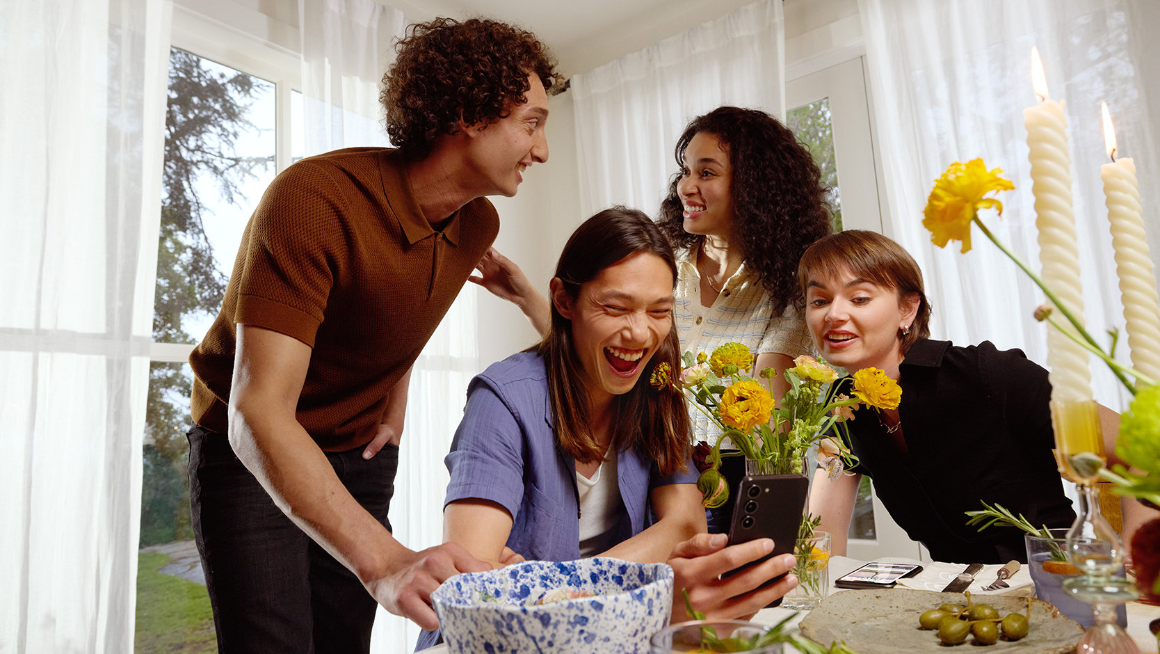 A group of friends at a dining table, gather around a phone.