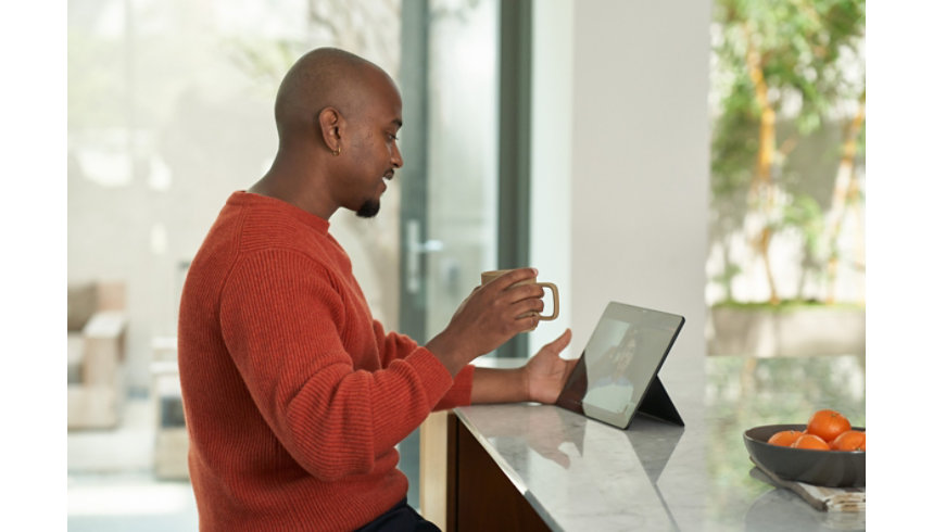 A smiling man in a red sweater sits in an open room holding a coffee mug and watching a tablet screen.