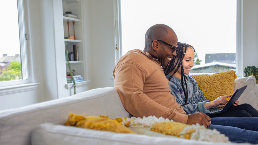 A father and daughter sitting next to each other on a sofa at home, looking at the screen of a laptop together and smiling