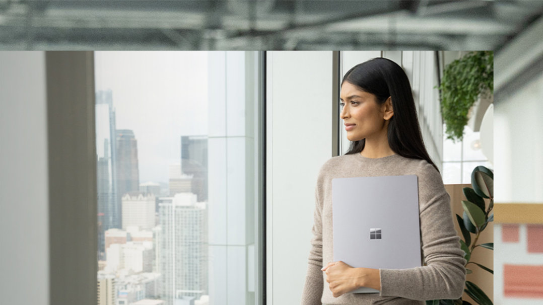 A person holds their Surface Laptop 5 device against their body with one arm while looking across the room