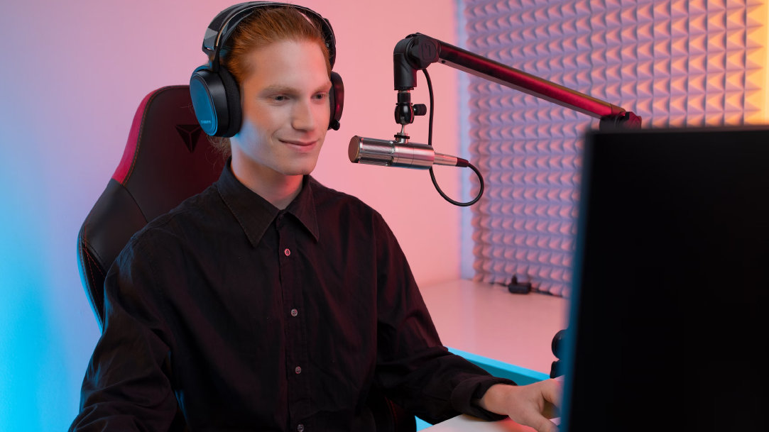 A person wearing headphones sits at a computer and microphone with a slight smile on their face