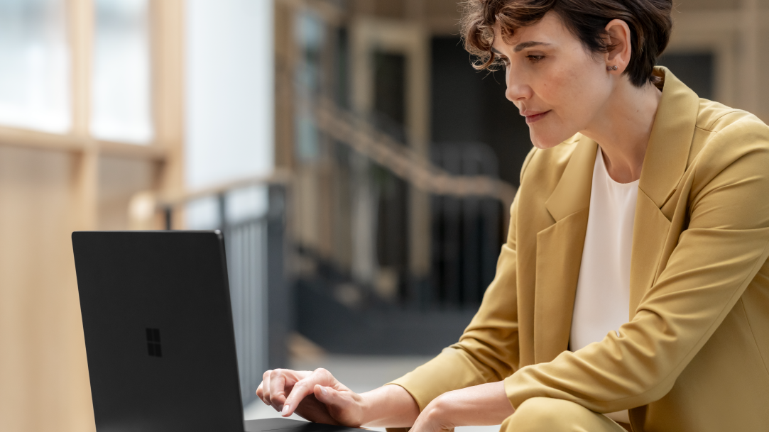 A woman in a yellow suit scrolls on a black Surface laptop