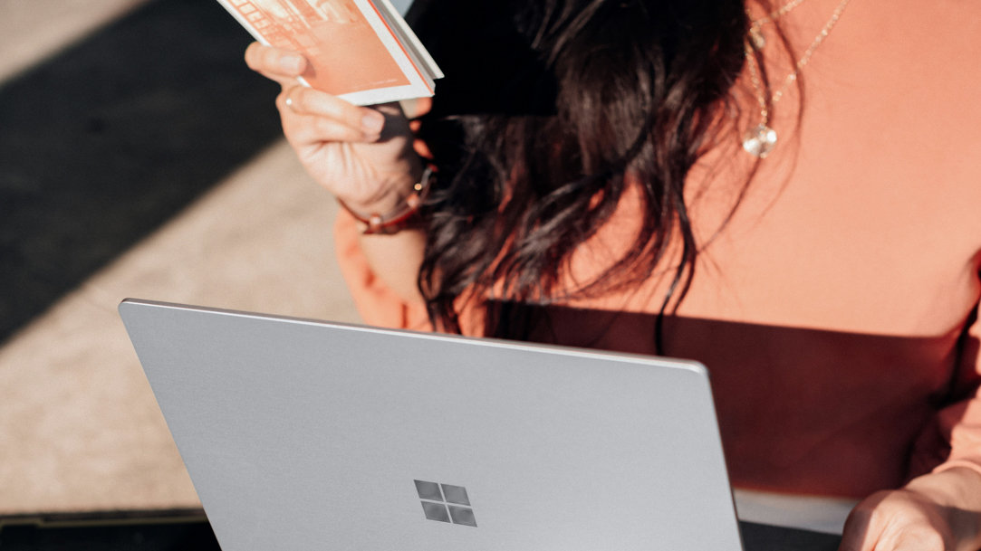 A woman in orange shirt and white pants holding a book and silver Microsoft Surface laptop computer