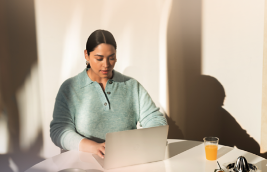 An adult female sits in a blue chair indoors and works on a Surface Laptop