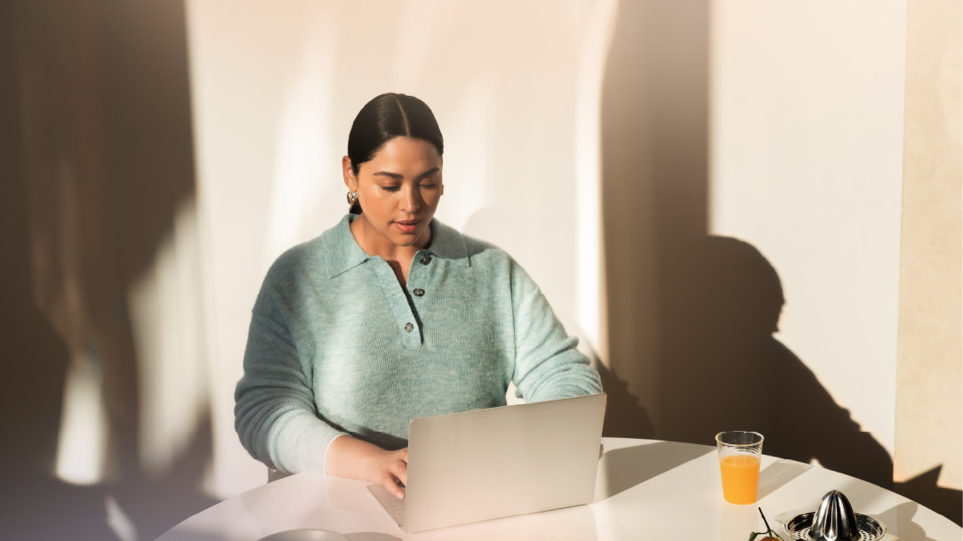 An adult female sits in a blue chair indoors and works on a Surface Laptop