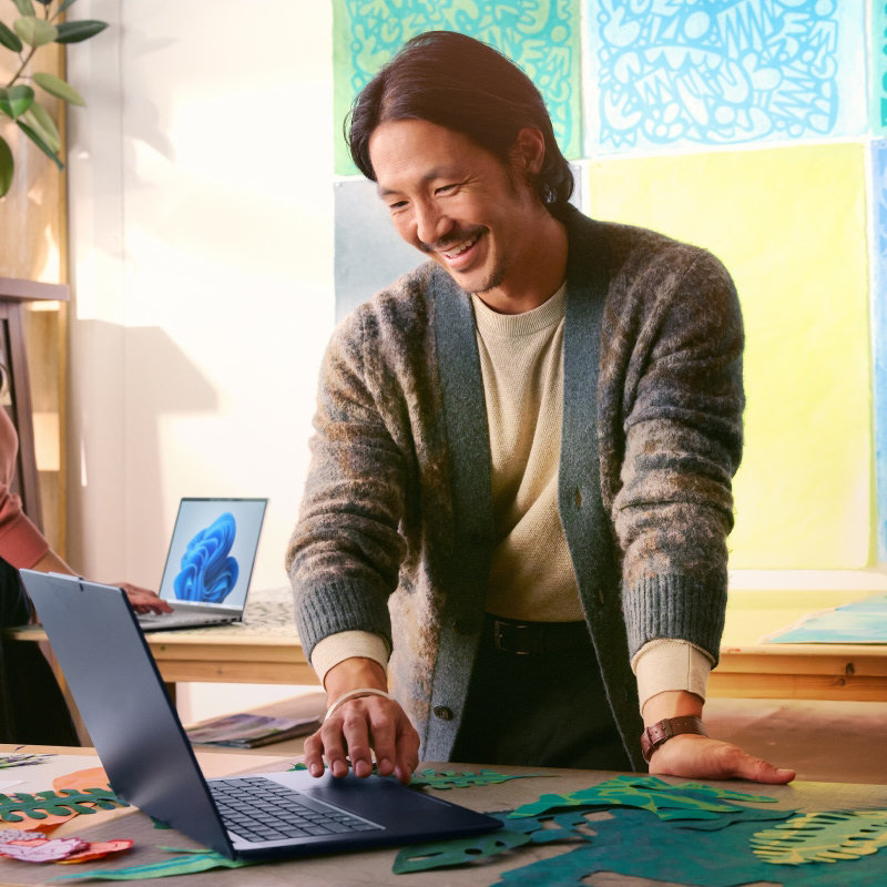 A smiling man standing at a desk and working on an open laptop with art prints in the background