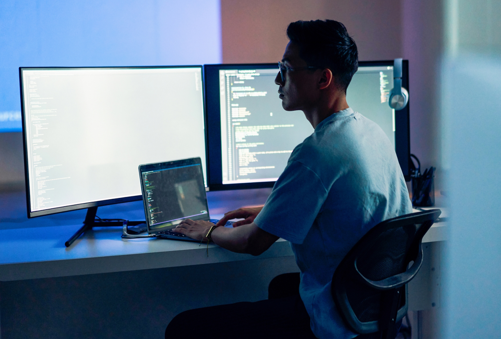 A man wearing glasses is working code at a 3 screen workstation.