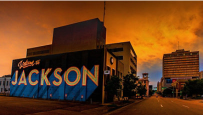 View of a downtown street with a vibrantly colored mural on the closest building reading “Welcome to Jackson”.