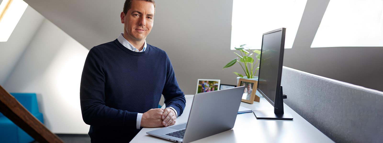 Man standing at office desk with open Windows laptop device on table and monitor off to the side