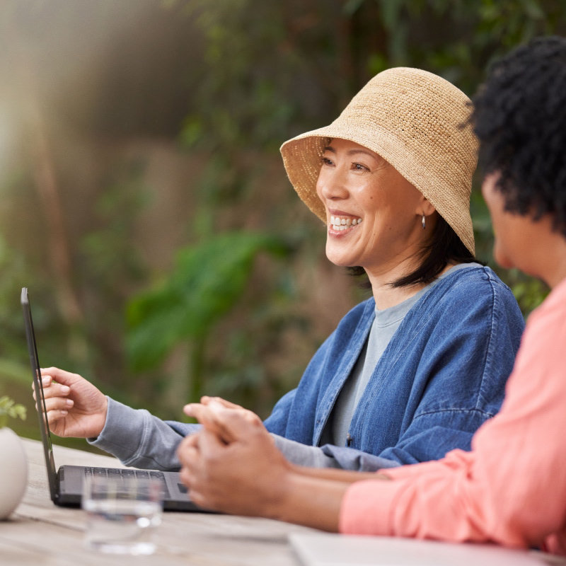 Two smiling women sitting at a table outside and looking at an open laptop