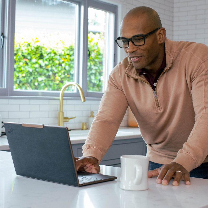 A smiling man standing at a kitchen counter working on an open laptop