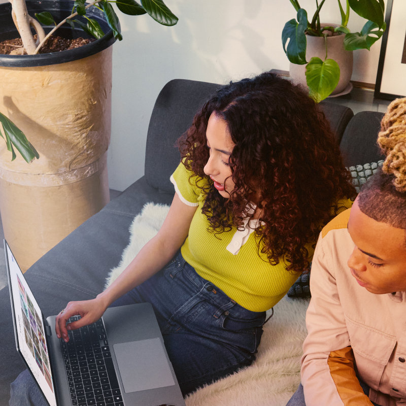 Two people sitting on a couch and looking at a laptop together with potted plants in the background