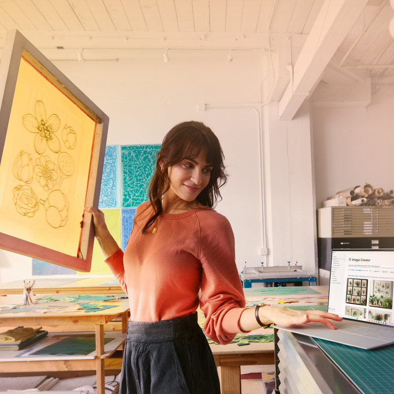 A woman in an art studio holding up a framed flower print with her right hand while looking at pictures on an open laptop to her left