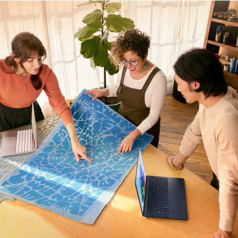 Three people, in an open, bright room with sheer curtained windows, plants, and a bookshelf in the background, are leaning over a table with two open laptops to look at a large blue abstract print.