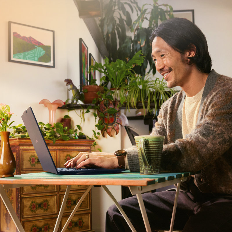 A man sitting at a table and working on a laptop with several plants, a dresser, and framed art prints in the background.