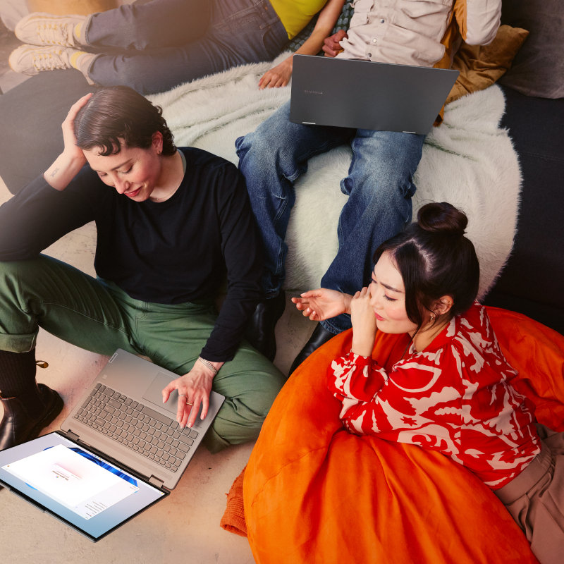 Four people are lounging in a living room. Two of the people are looking at an open laptop while sitting on the floor and lying on a beanbag and the other two are looking at another open laptop while lounging on the couch.