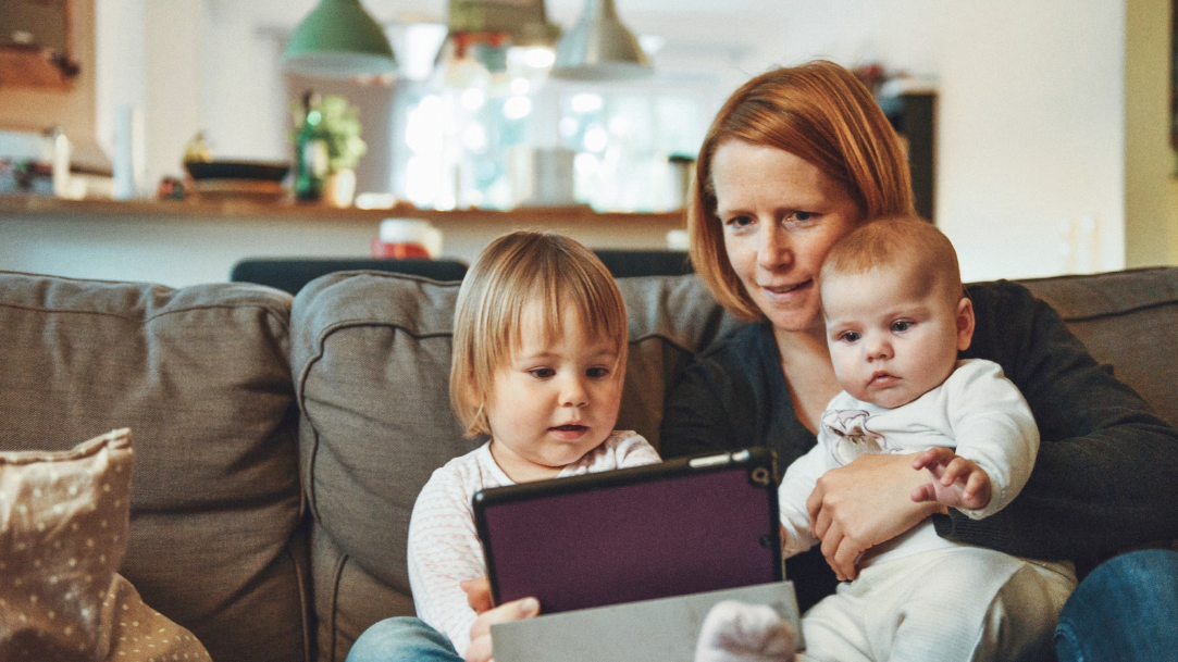 Two babies and woman sitting on sofa while holding baby and watching on tablet