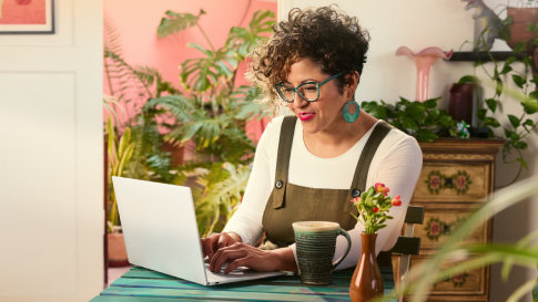 A smiling woman sitting at a blue table working on an open laptop with a dresser, plants, and an art print in the background.