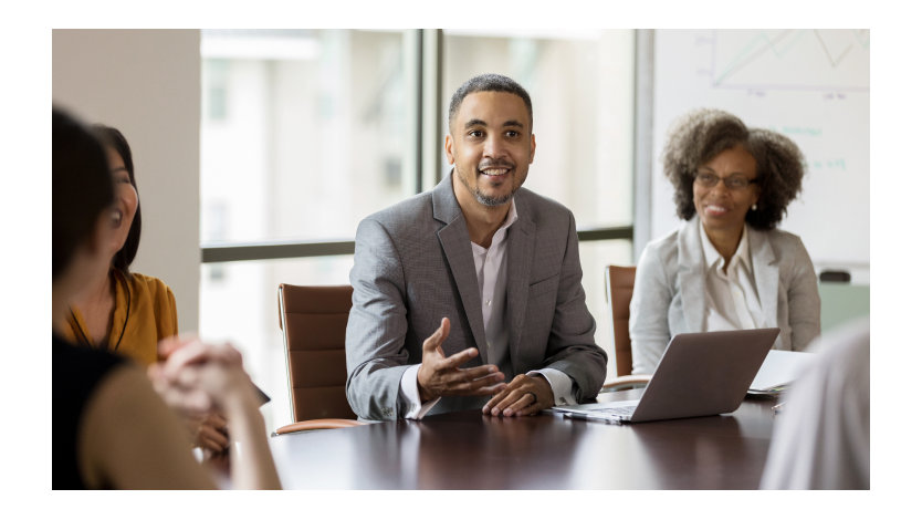 Person in boardroom speaking to others while using a laptop