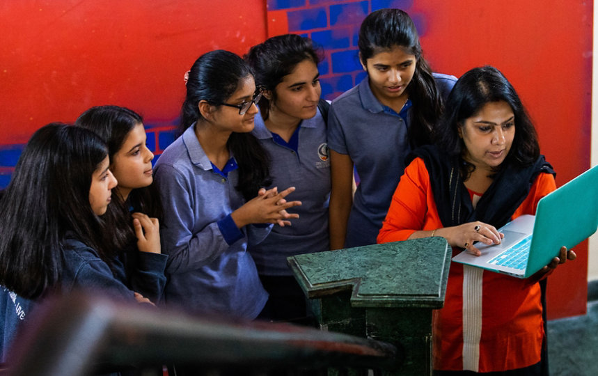 A teacher informally presenting with a laptop to several girls in a colorful hallway.