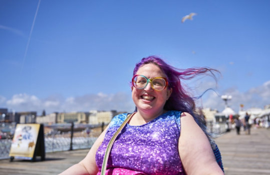 A smiling person with purple hair and colorful glasses enjoys a sunny day outdoors.