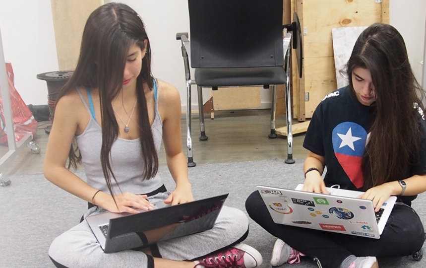 Two female students sitting cross-legged on the floor and working on laptops in a CS class