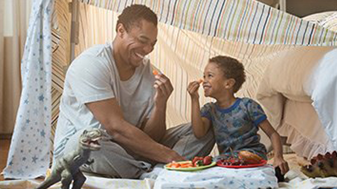 Laughing father and son eating in blanket fort while playing together in a bedroom