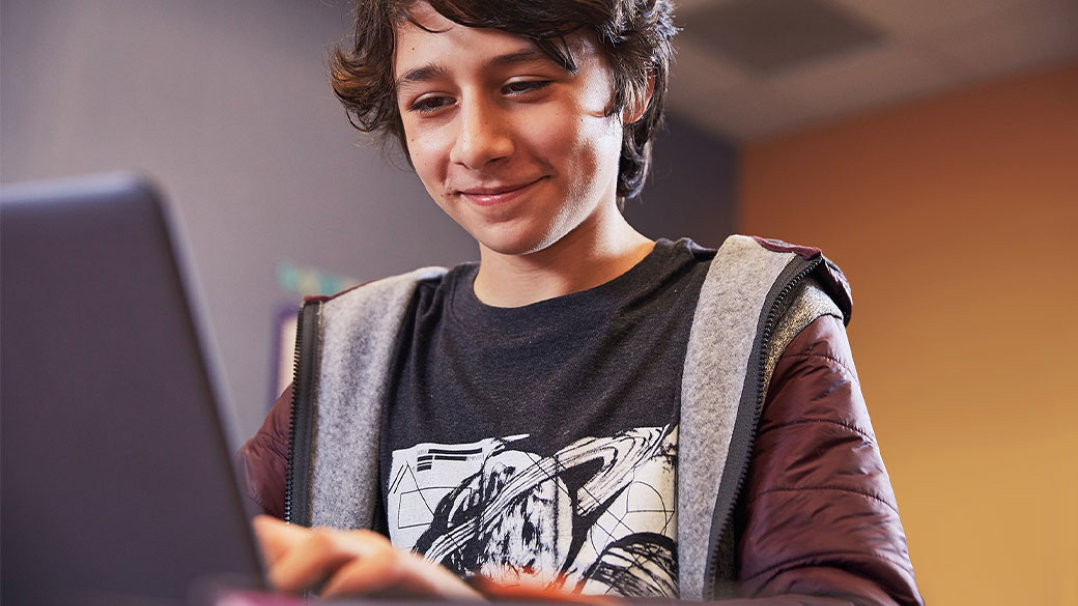 Male middle school student grins while using laptop, seated at desk in bedroom