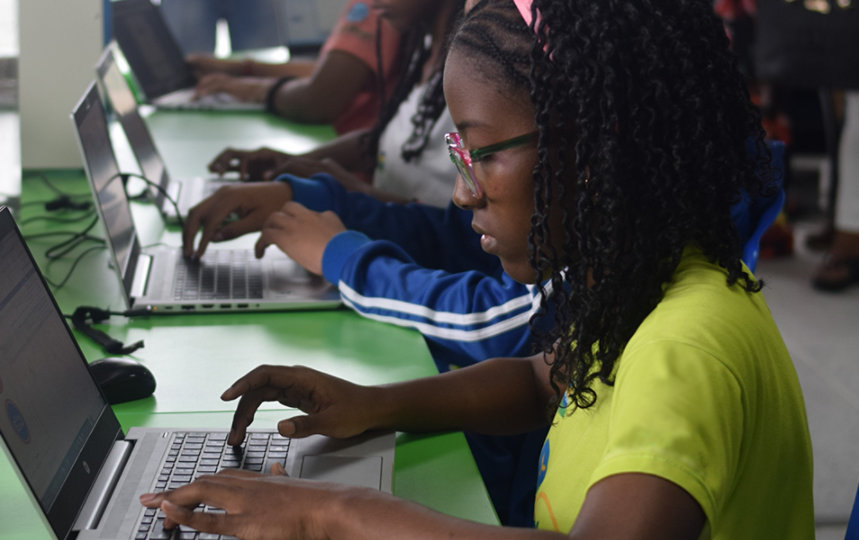 A student in the center is working on her laptop, and more students are in the background in a computer lab.