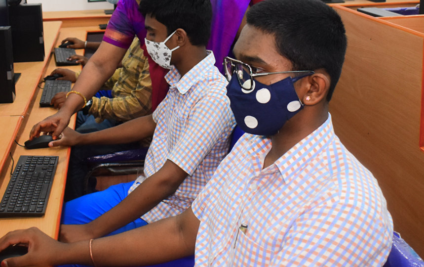 A young man wearing a mask and using a mouse sits in front of a computer inside a computer lab with other students.