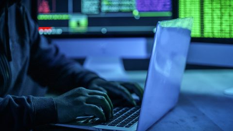 Person in a hoodie typing on a laptop, surrounded by computer screens with colorful code in a dimly lit room.