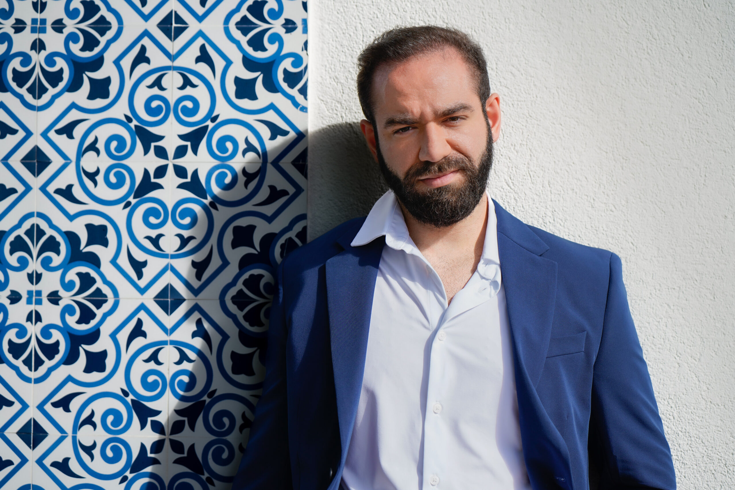 A man in a blue suit leaning against a tiled wall.