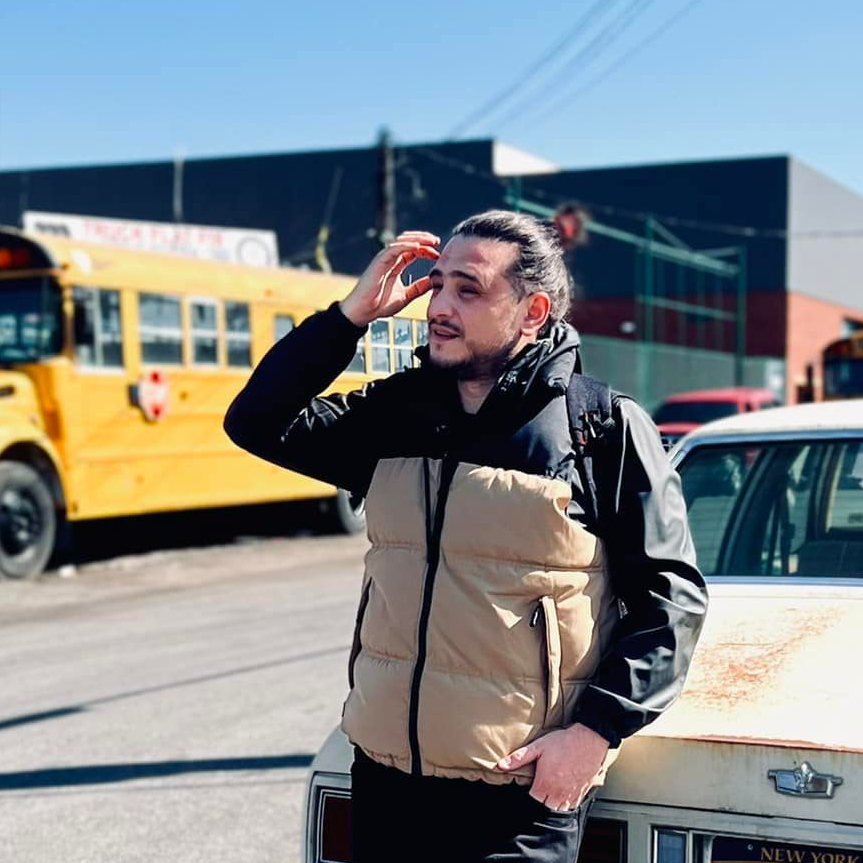 Man in a black jacket and beige vest standing by an old car with a yellow school bus in the background on a sunny day.