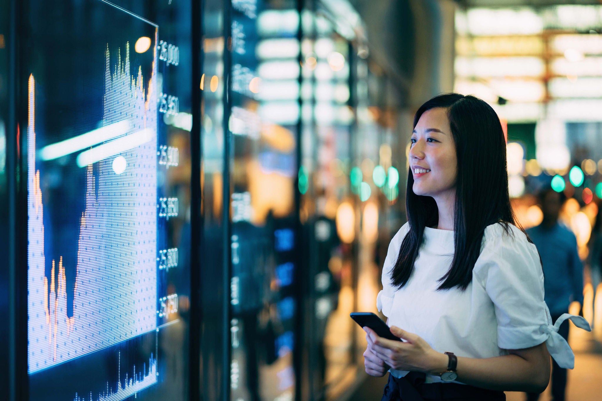 Confidence young Asian businesswoman checking financial trading data on smartphone by the stock exchange market display screen board in downtown financial district