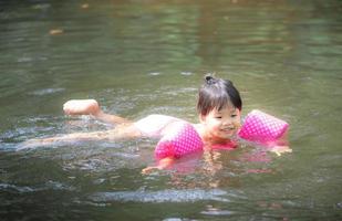 Little Asian girl wearing inflatable sleeves swimming in nature water on holiday photo