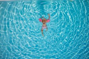 Cute little toddler girl swimming in turquoise pool with inflatable arms aids support, top view. photo