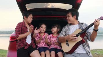 família feliz aproveitando a viagem nas férias de verão. mãe e filho sentam-se no porta-malas do carro cantando junto com o pai tocando violão. conceito de família de férias e viagens. video